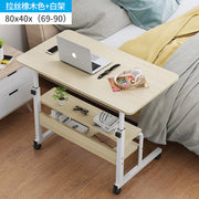 Light wood mobile laptop desk with wheels and storage shelves next to a bed