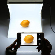 Person using a smartphone to photograph a lemon inside a professional white light box studio.