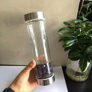 Hand holding a clear glass water bottle with purple crystals at the bottom