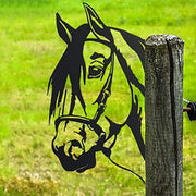 Black silhouette horse head metal art mounted on a wooden fence post in a green field.